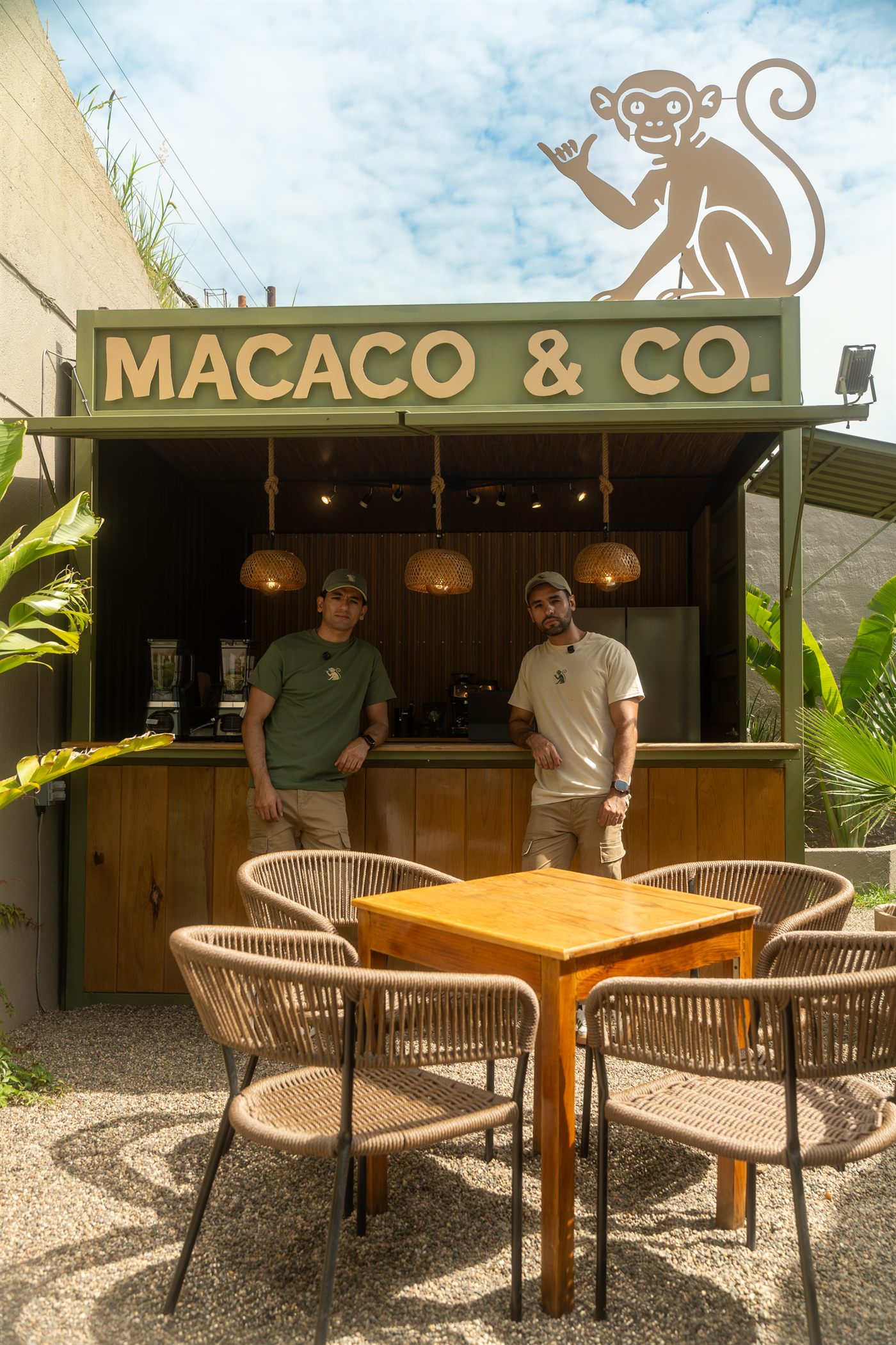 Los fundadores de Macaco & Co. recargados en la barra del local, con el letrero y el chango en el techo